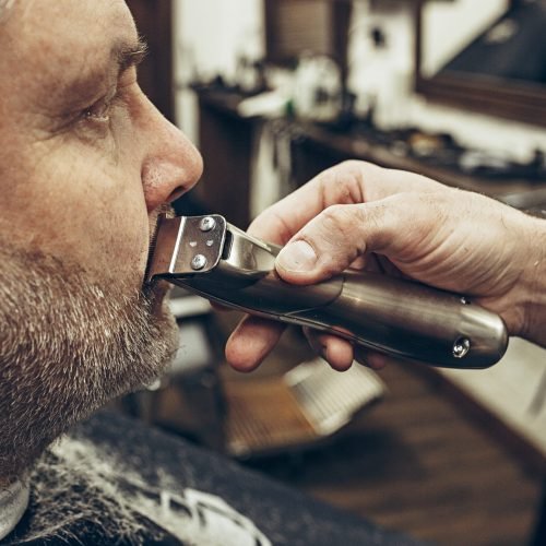 Close-up side profile portrait of handsome senior bearded caucasian man getting beard grooming in modern barbershop. Hairdresser serving client, making beard haircut using shaving machine. Barber shop concept.