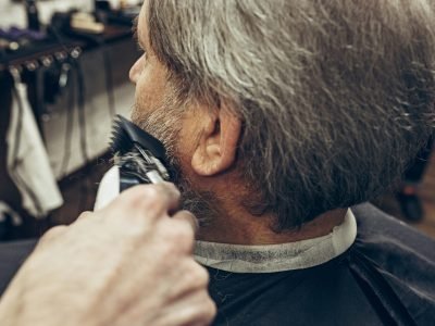 Close-up side back view handsome senior bearded caucasian man getting beard grooming in modern barbershop. Hairdresser serving client, making beard haircut using shaving machine. Barber shop concept.